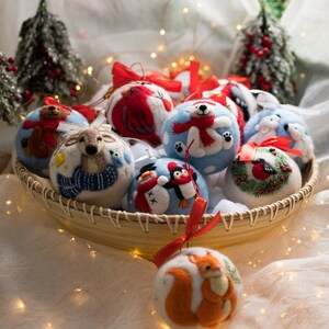 May include: A collection of round, felted wool Christmas ornaments featuring needle-felted designs of animals including a bear, deer, polar bear, penguin, snowman, squirrel, and birds, each topped with a ribbon loop for hanging in a woven basket.