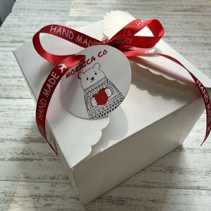 May include: A square white cardboard box tied with a red satin ribbon printed with the words HAND MADE. A round white tag is attached to the ribbon, featuring a black line drawing of a bear wearing a patterned sweater and holding a red heart.