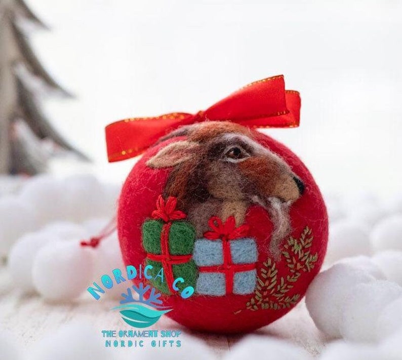 May include: A round red felt ornament featuring a needle-felted brown goat head, a green gift box, and a light blue gift box with red ribbons. A red satin ribbon bow is attached to the top. The text NORDICA CO THE ORNAMENT SHOP NORDIC GIFTS is visible.