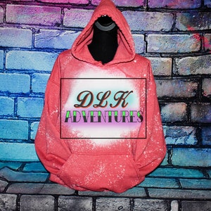 Puede incluir: Una sudadera con capucha de color rosa coral con un diseño de blanqueador blanco que dice "DLK ADVENTURES" en letras verdes y moradas.