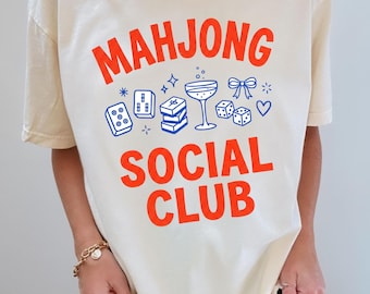 May include: An ivory t-shirt featuring the orange text "MAHJONG SOCIAL CLUB" printed on the front. Below the text are blue line-art graphics of mahjong tiles, a stacked set of tiles, a cocktail glass, a bow, and dice, accented with small stars.