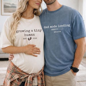 May include: A couple wearing matching t-shirts. One shirt is ivory and says "growing a tiny human" with a due date of November 2026. The other shirt is blue and says "dad mode loading" with "EST.2006".