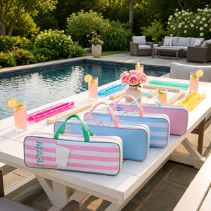 May include: Three rectangular zipped carrying cases for mahjong tiles, arranged in a row on a white outdoor table. The cases feature striped patterns in pink and white, light blue and white, and solid pink, each with top handles and the word MAHJ.