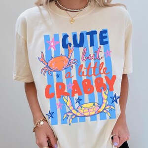 May include: Ivory-colored t-shirt with the text "Cute but a little Crabby" in blue, pink, and orange. The shirt features two crab illustrations and blue and pink star accents. The shirt has short sleeves.