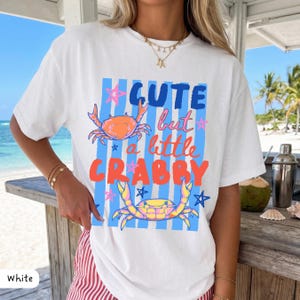 May include: White t-shirt with a graphic design featuring the text "Cute but a little Crabby" in blue and orange. The design includes illustrations of crabs and stars on a blue striped background. A casual, beach-themed apparel item.