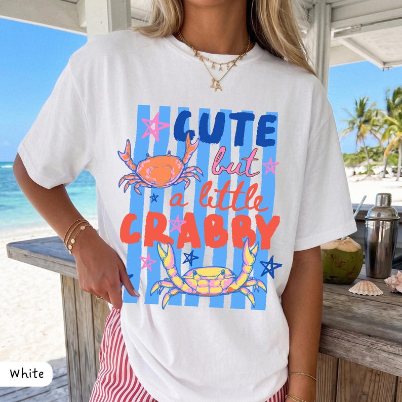 May include: White t-shirt with a graphic design featuring the text "Cute but a little Crabby" in blue and orange. The design includes illustrations of crabs and stars on a blue striped background. A casual, beach-themed apparel item.