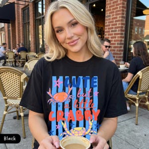May include: Black t-shirt with the text "Cute but a little Crabby" in blue, orange, and pink. The design includes two crab illustrations. The shirt is worn by a person sitting outdoors.