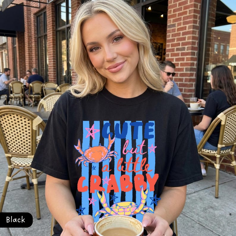 May include: Black t-shirt with the text "Cute but a little Crabby" in blue, orange, and pink. The design includes two crab illustrations. The shirt is worn by a person sitting outdoors.