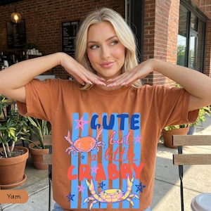 May include: A brown t-shirt with the text "CUTE but a little CRABBY" in blue, pink, and orange. The shirt features two crab illustrations and blue vertical stripes. The word "Yam" is at the bottom left.