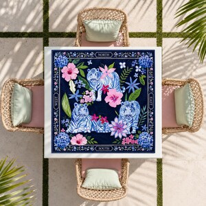 May include: A square tablecloth featuring a dark blue background with three white tigers, pink hibiscus flowers, and blue hydrangea clusters. The border includes the words NORTH, SOUTH, EAST, and WEST printed in white text on a decorative frame.