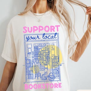 May include: An ivory short-sleeve t-shirt featuring a blue line-art illustration of a bookstore interior with a cat, a chair, and stacks of books. The text "SUPPORT YOUR LOCAL BOOKSTORE" is printed in pink block letters above and below the graphic.