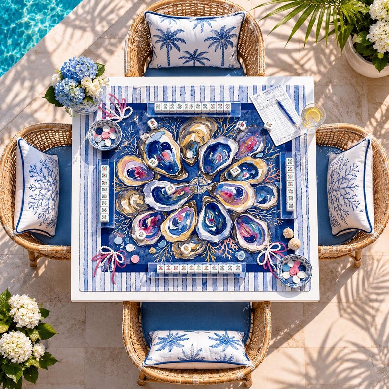 May include: A square game table featuring a central mat with a vibrant oyster shell illustration in shades of blue, gold, and pink. The table is surrounded by four wicker chairs with blue and white palm and coral patterned cushions and pillows.