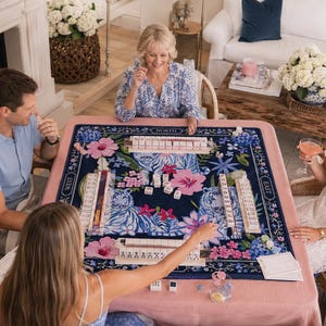 May include: A square mahjong game set featuring a dark blue playing mat with a vibrant floral pattern in shades of pink, blue, and white. The mat includes labelled directional markers for North, South, East, and West, with four tile racks and white tiles.