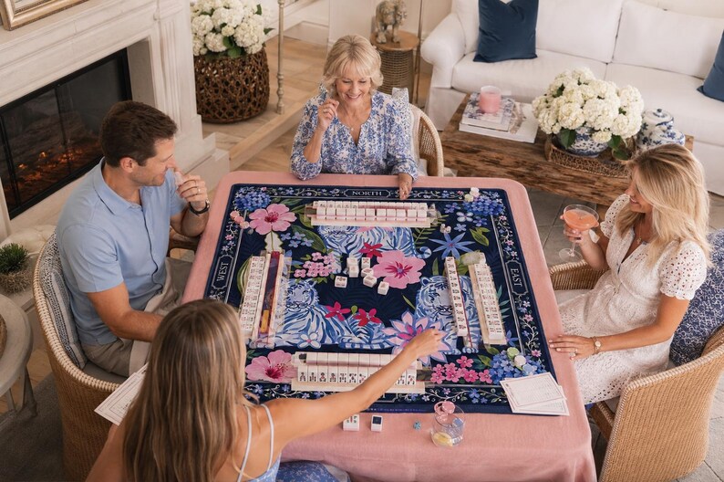 May include: A square mahjong game set featuring a dark blue playing mat with a vibrant floral pattern in shades of pink, blue, and white. The mat includes labelled directional markers for North, South, East, and West, with four tile racks and white tiles.