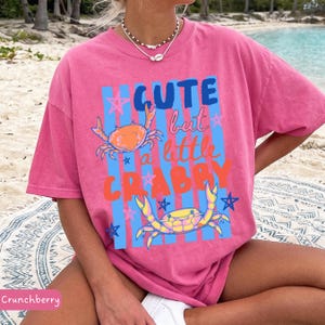May include: Pink t-shirt with the text "Cute but a little Crabby" in blue, orange, and red. The shirt features two crab illustrations and blue star accents. The shirt is a relaxed fit.
