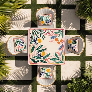 Op de afbeelding: Een vierkante buitentafel met een cr&egrave;mekleurig blad voorzien van een botanisch patroon van groene bladeren, oranje citrusvruchten en roze bessen, omringd door vier witte gevlochten stoelen, elk met een bijpassend vierkant sierkussen.