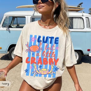 May include: Ivory-colored t-shirt with the text "Cute but a little Crabby" in blue and orange. The shirt features crab illustrations and blue vertical stripes. The shirt is a relaxed fit.