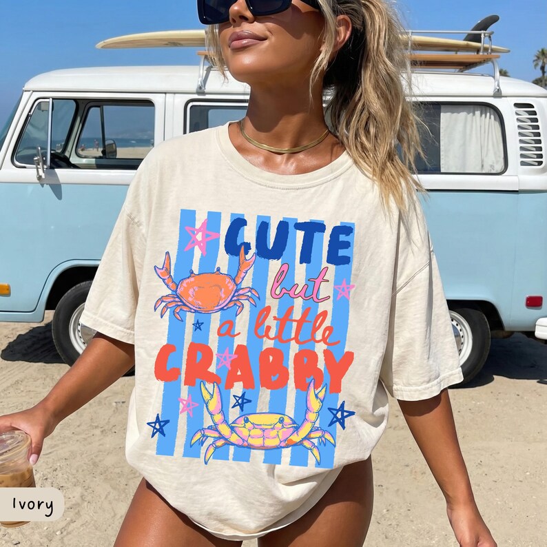 May include: Ivory-colored t-shirt with the text "Cute but a little Crabby" in blue and orange. The shirt features crab illustrations and blue vertical stripes. The shirt is a relaxed fit.