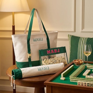 May include: A cream and dark green canvas tote bag with the text MAHJ, a matching cylindrical drawstring bag with the text MAHJ, and a clear zippered pouch containing mahjong tiles with the pink embroidered text MAHJ on the front.