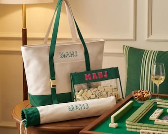 May include: A cream and dark green canvas tote bag with the text MAHJ, a matching cylindrical drawstring bag with the text MAHJ, and a clear zippered pouch containing mahjong tiles with the pink embroidered text MAHJ on the front.