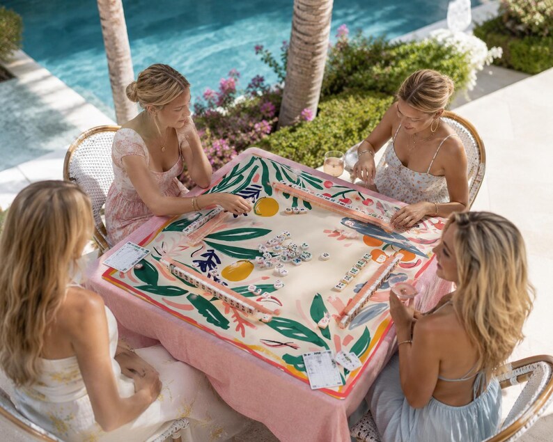 Op de afbeelding: Een vierkante mahjongtafel bedekt met een cr&egrave;mekleurig kleed met een levendige botanische print van groene bladeren, roze bloemen en gele citrusvruchten. De tafel is ingericht met vier houten tegelhouders en een verzameling witte mahjongtegels.