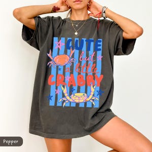 May include: Dark gray t-shirt with the text "Cute but a little Crabby" in blue, pink, and red. The shirt features illustrations of crabs and stars on a blue striped background. A casual, oversized tee.
