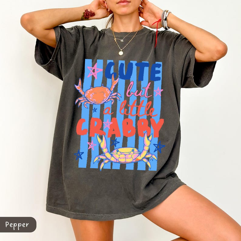 May include: Dark gray t-shirt with the text "Cute but a little Crabby" in blue, pink, and red. The shirt features illustrations of crabs and stars on a blue striped background. A casual, oversized tee.