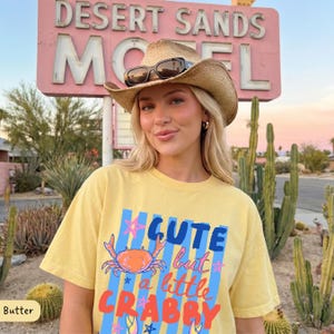 May include: A woman wearing a straw cowboy hat and sunglasses, and a yellow t-shirt with the text "Cute but a little Crabby" and a crab graphic. The background includes a pink motel sign and desert plants.