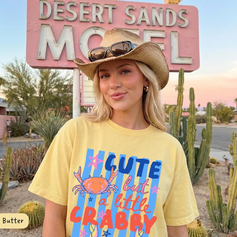 May include: A woman wearing a straw cowboy hat and sunglasses, and a yellow t-shirt with the text "Cute but a little Crabby" and a crab graphic. The background includes a pink motel sign and desert plants.