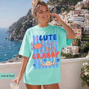 May include: A light turquoise t-shirt with the text "Cute but a little Crabby" in a colorful design. The shirt features two crab illustrations and star accents. The shirt is a relaxed fit.