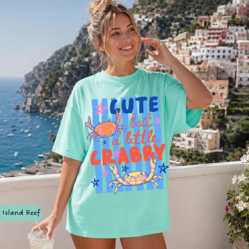 May include: A light turquoise t-shirt with the text "Cute but a little Crabby" in a colorful design. The shirt features two crab illustrations and star accents. The shirt is a relaxed fit.