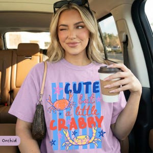 May include: A light purple t-shirt with the text "CUTE but a little CRABBY" in blue, orange, and red. The shirt features two crab illustrations and blue star accents. The shirt is being worn.