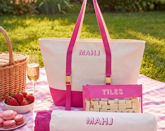 May include: A cream and hot pink canvas tote bag with the text MAHJ, a matching cylindrical case with the text MAHJ, and a clear zippered pouch with the text TILES containing white rectangular game pieces, all featuring hot pink straps and gold buckles.