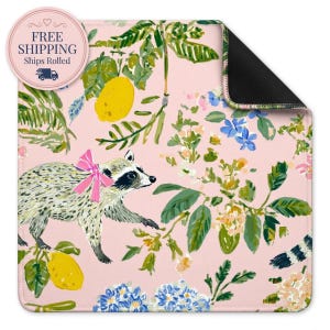 May include: A rectangular mat with a light pink background featuring a repeating pattern of a raccoon wearing a bright pink bow, yellow lemons, green foliage, and clusters of small blue and white flowers. The mat has a black underside.