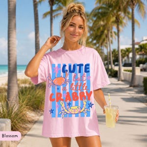 May include: A light pink t-shirt with the text "Cute but a little Crabby" in blue, orange, and red. The shirt features crab illustrations and blue stripes. The model is holding a beverage with a straw.