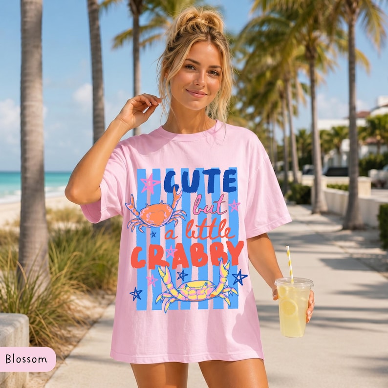 May include: A light pink t-shirt with the text "Cute but a little Crabby" in blue, orange, and red. The shirt features crab illustrations and blue stripes. The model is holding a beverage with a straw.