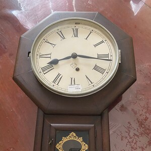 May include: An antique wooden wall clock with a white face and black Roman numeral hour markers. The clock has a small glass window with a decorative gold frame.