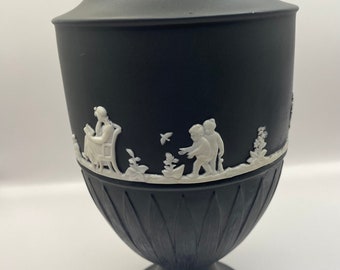 May include: Black and white ceramic vase with a white relief design of figures and flowers. The vase has a fluted body and a round base.