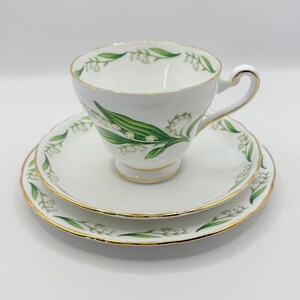 May include: A white teacup and saucer set with a floral design. The teacup and saucer have a gold trim and a green and white lily of the valley pattern. The set is on a white background.