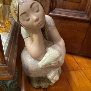 Puede incluir: Figura de cerámica de una persona sentada con los ojos cerrados, apoyando la cabeza en una mano. La figura tiene un tono de piel claro, cabello rubio y un vestido azul claro y blanco. La figura sostiene un objeto blanco.