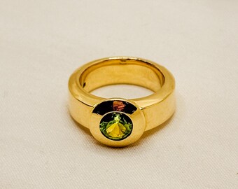 May include: A gold ring with a green gemstone set in a circular bezel. The ring has a thick band.