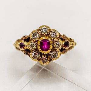 Puede incluir: Un anillo de oro con un rubí rojo central de corte ovalado rodeado por un grupo de pequeños diamantes blancos redondos. Dos rubíes rojos adicionales están engastados en los lados de la banda de oro texturizada con un acabado grabado detallado.