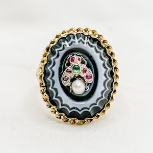 May include: A gold ring featuring a black and white agate stone set in a gold bezel. The stone is encircled by a decorative gold border. The centre of the stone showcases a small pearl and a cluster of red and green gemstones.
