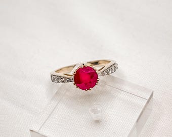 Pode incluir: Um anel de ouro com uma grande gema de rubi vermelha redonda cercada por pequenos diamantes transparentes.