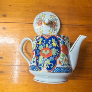May include: White ceramic teapot with a lid, featuring a blue, red, and yellow floral design. The teapot has a curved handle and spout, with gold trim. The lid is decorated with similar floral patterns.