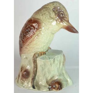 May include: A ceramic kookaburra bird figurine perched on a tree stump. The bird has brown and cream-colored feathers, a long beak, and is looking down. The tree stump is light green with brown accents.