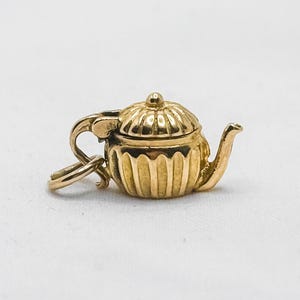 May include: A small, gold-colored teapot charm with a lid and spout. The charm has a loop for attaching to a bracelet or necklace. The teapot is detailed with vertical lines and a curved handle.