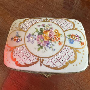 Puede incluir: Una caja de porcelana blanca para alhajas con un diseño floral y detalles dorados. La tapa presenta un ramo floral rosa, amarillo y blanco sobre un fondo a cuadros rosa y blanco.