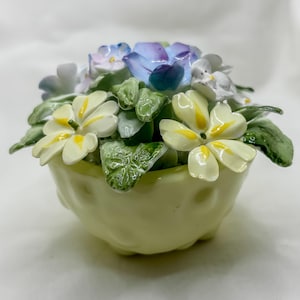 May include: A ceramic floral arrangement in a pale yellow pot. The arrangement features a variety of flowers in shades of blue, white, and yellow, with green leaves. The pot has a rounded shape and sits on three small feet.