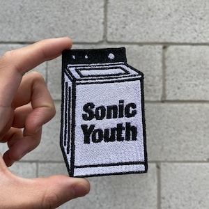 May include: Embroidered patch featuring a stylized washing machine design. The patch is primarily white with black detailing and the text "Sonic Youth" in a bold, sans-serif font. The patch is held up by a hand.